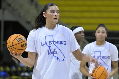 لاعبة كرة سلة من جامعة كاليفورنيا في لوس أنجلوس ترتدي قميصًا يحمل عبارة \"LA STRONG\" وتحمل كرة سلة، مع زميلتين في الخلفية.
