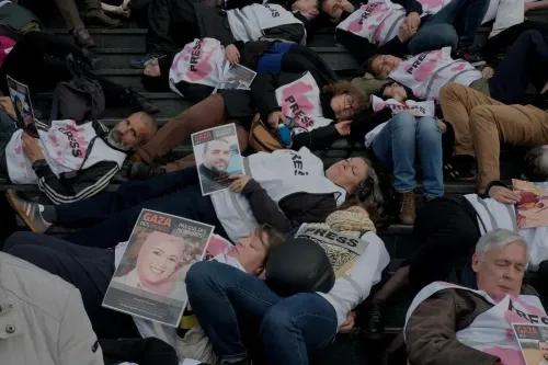 احتجاج لصحفيين فرنسيين في باريس، مستلقين على السلالم، يرتدون سترات تحمل شعار "PRESS" وبقع حمراء، يحملون لافتات تحمل صور زملائهم القتلى.