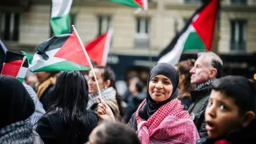 امرأة ترتدي الحجاب وتبتسم بينما تحمل علم فلسطين بين حشد من المتظاهرين، تعبيرًا عن التضامن مع القضية الفلسطينية.