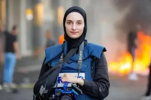 صحفية فلسطينية ترتدي سترة صحفية زرقاء وتحمل كاميرا، تقف أمام خلفية مشتعلة، تعكس التوترات في القدس المحتلة.