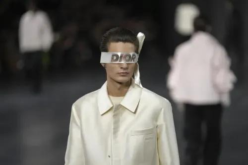 عارض أزياء يرتدي قميصًا كريميًا مع نظارات تحمل شعار "DIOR"، بينما يسير في عرض ديور خلال أسبوع الموضة في باريس.