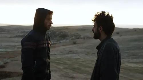 مشهد من فيلم \"لا أرض سواها\" يظهر شابين فلسطينيين يتحدثان في منطقة مفتوحة بالضفة الغربية، مع خلفية طبيعية تعكس التوترات المستمرة.