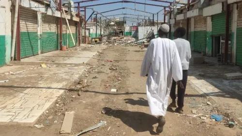شارع مدمّر في قرية سودانية، يظهر فيه رجلان يسيران وسط الأنقاض والمخلفات، مما يعكس آثار النزاع المستمر في البلاد.