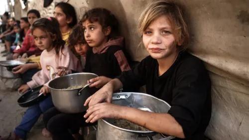 أطفال يجلسون في صف، يحملون أواني طعام، يعكسون معاناة السكان في غزة بسبب نقص المساعدات الإنسانية في ظل الحصار.
