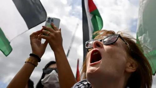 امرأة تتظاهر في احتجاج لدعم فلسطين، ترفع هاتفها وتصرخ بحماس، مع أعلام فلسطينية في الخلفية.