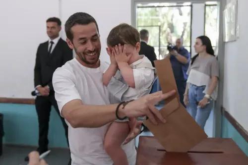 مواطن في أوروجواي يحمل طفلاً أثناء إدلائه بصوته في الانتخابات، مع وجود صندوق الاقتراع في المقدمة وأشخاص آخرين في الخلفية.