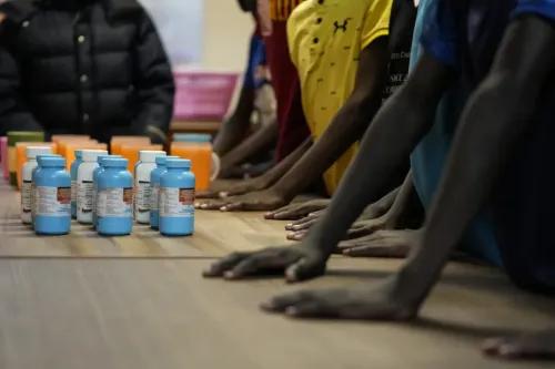 أيدي أطفال في دار نيومباني للأطفال تمتد نحو زجاجات الأدوية المضادة للفيروسات القهقرية، تعبير عن الأمل في حياة أفضل.