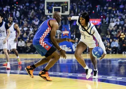 لاعبة كرة سلة تت dribble الكرة في مباراة، بينما يتفاعل لاعب آخر معها، مع جمهور خلفهم في ملعب مزدحم.