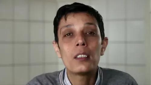 مازن الحمادة، الناشط السوري، يتحدث بوجه متألم في مكان مغلق، مع خلفية جدران بيضاء، معبراً عن معاناته في سجن صيدنايا.